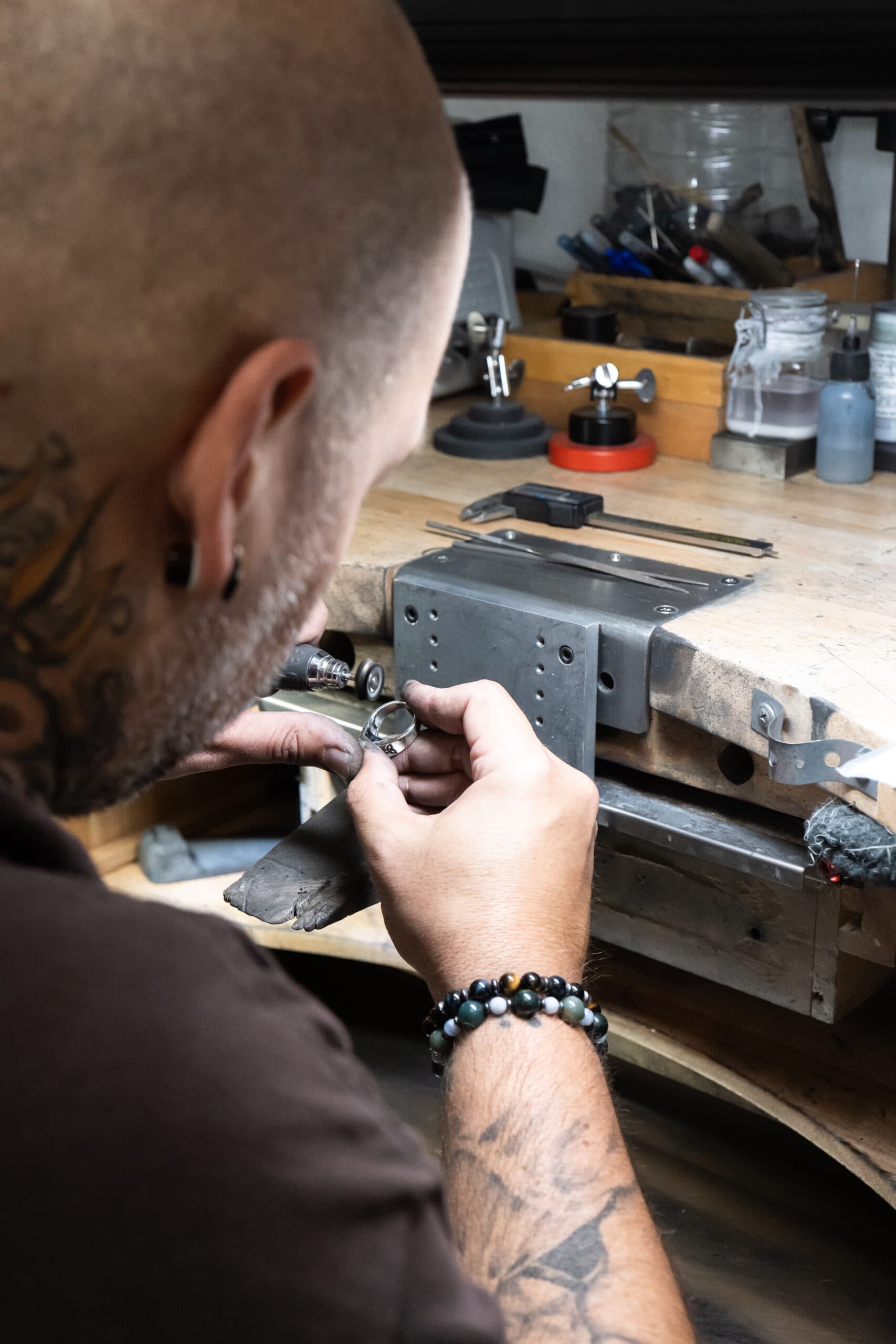 Tattooed jeweler with bead bracelet hand-finishing a silver ring at a workbench