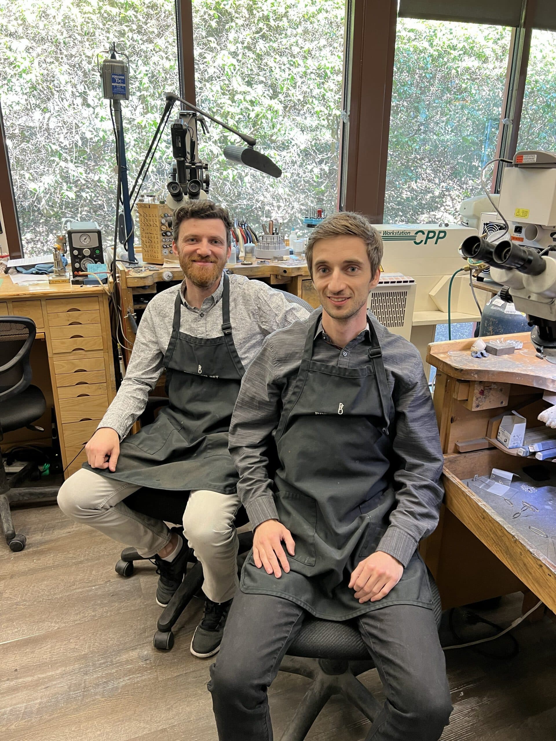 Two smiling jewelers in dark aprons seated at a jewelry workshop bench