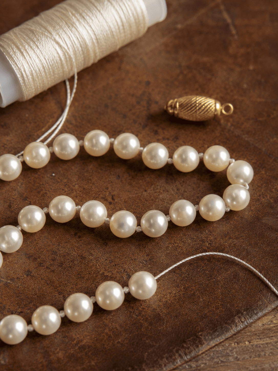 Knotted pearl necklace strand mid-restringing beside ivory silk thread spool and gold barrel clasp