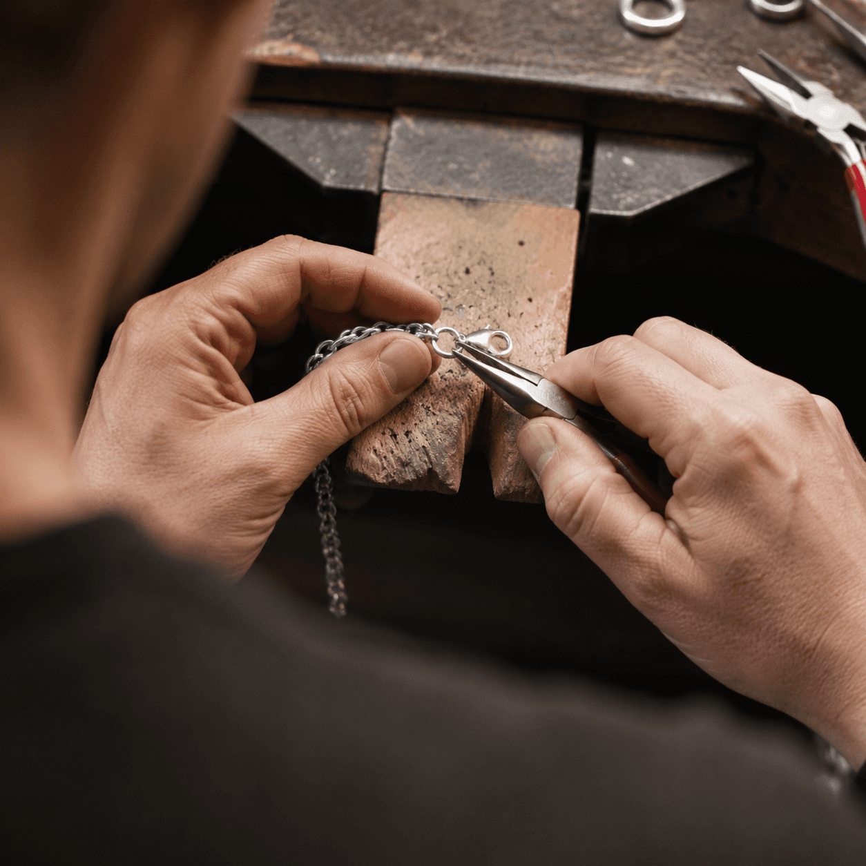 A jeweler using fine-tipped pliers to attach a new lobster-claw clasp to a silver bracelet, overhead shot on a wooden