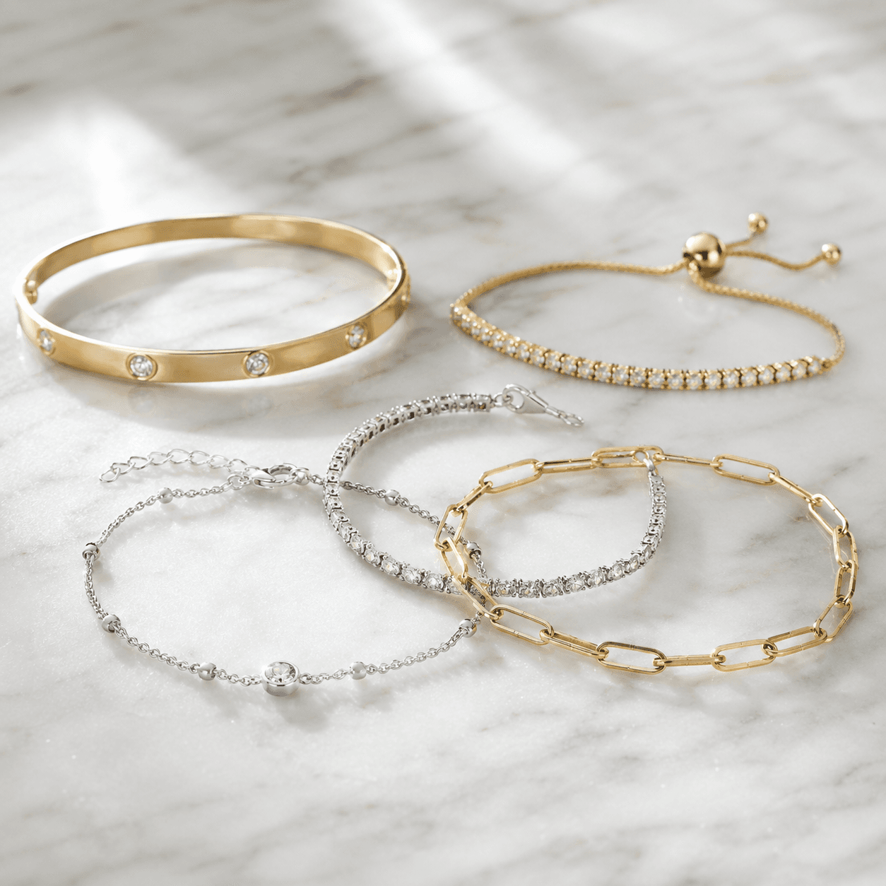 A collection of gold and silver bracelets laid flat on a white marble surface, including a tennis bracelet and a bangle