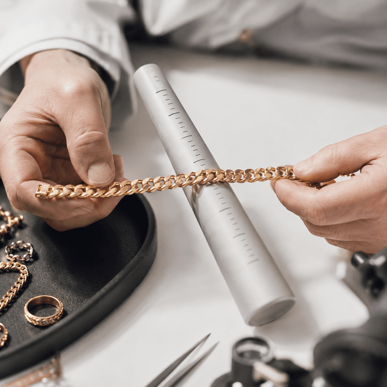 A jeweler's hands carefully measuring a gold chain bracelet against a sizing gauge on a clean white workbench, soft overhead