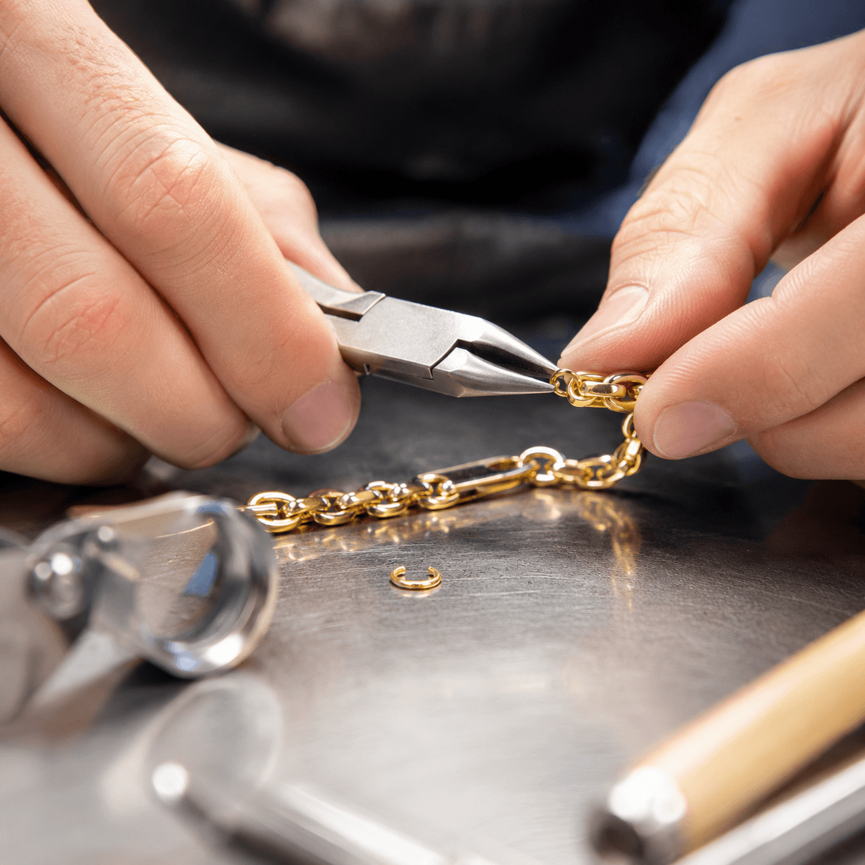 A close-up of a jeweler using fine pliers to remove a link from a yellow gold bracelet on a polished steel workbench, bright
