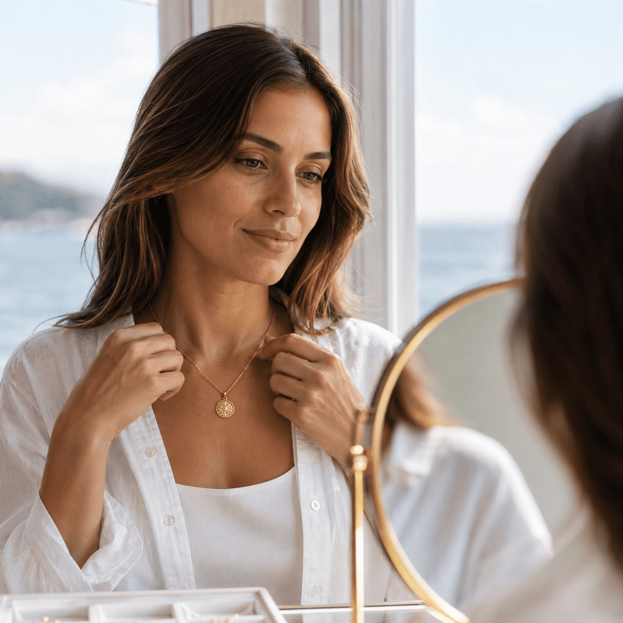 A woman standing near a sunlit window trying on a gold pendant necklace in front of a small mirror, natural coastal light