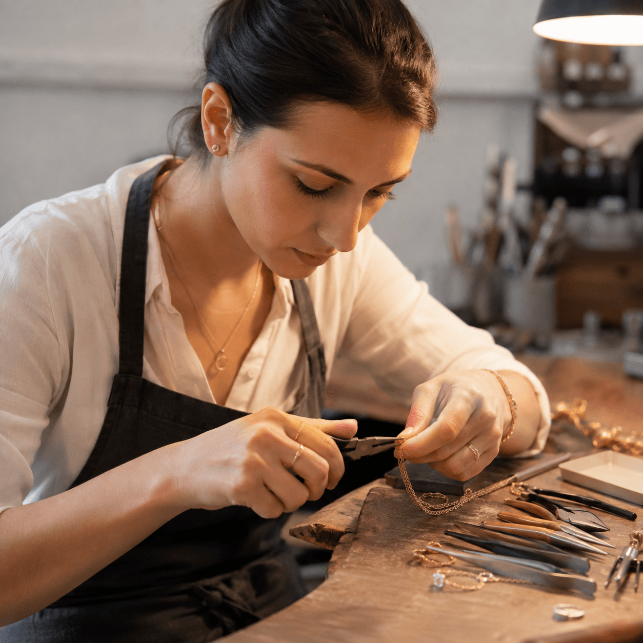 A female jeweler using fine-tip pliers to adjust a gold chain on a workbench, overhead warm workshop lighting, tools neatly