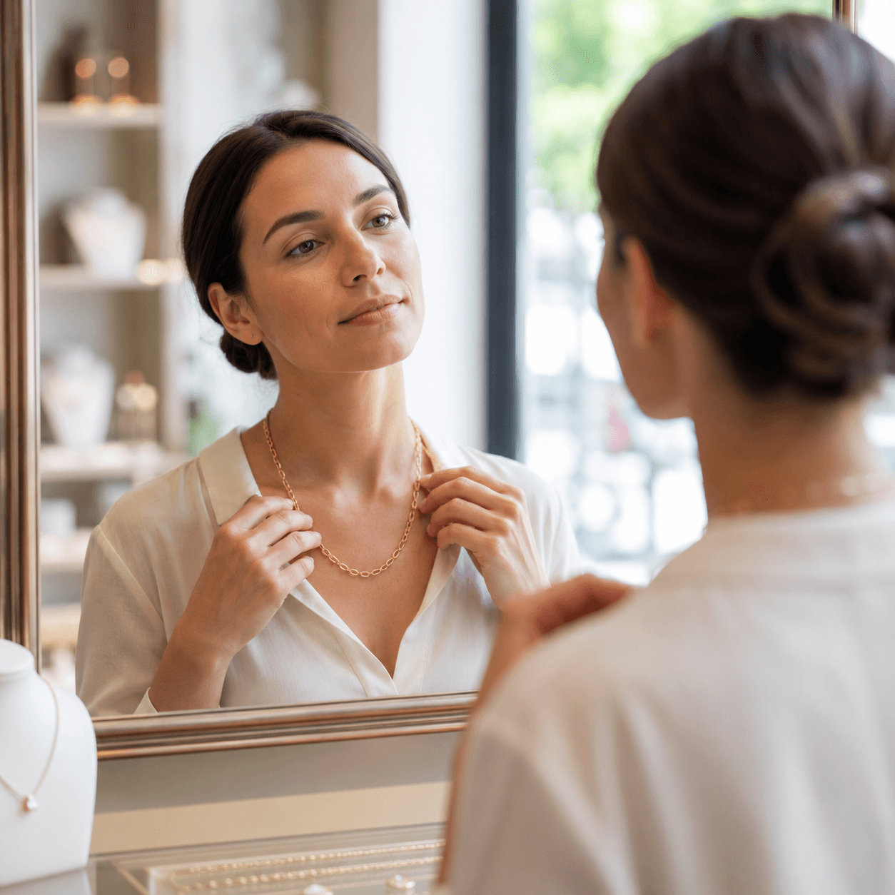 An elegant woman in a neutral blouse examining the drape of a gold chain necklace in a mirror inside a softly lit jewelry