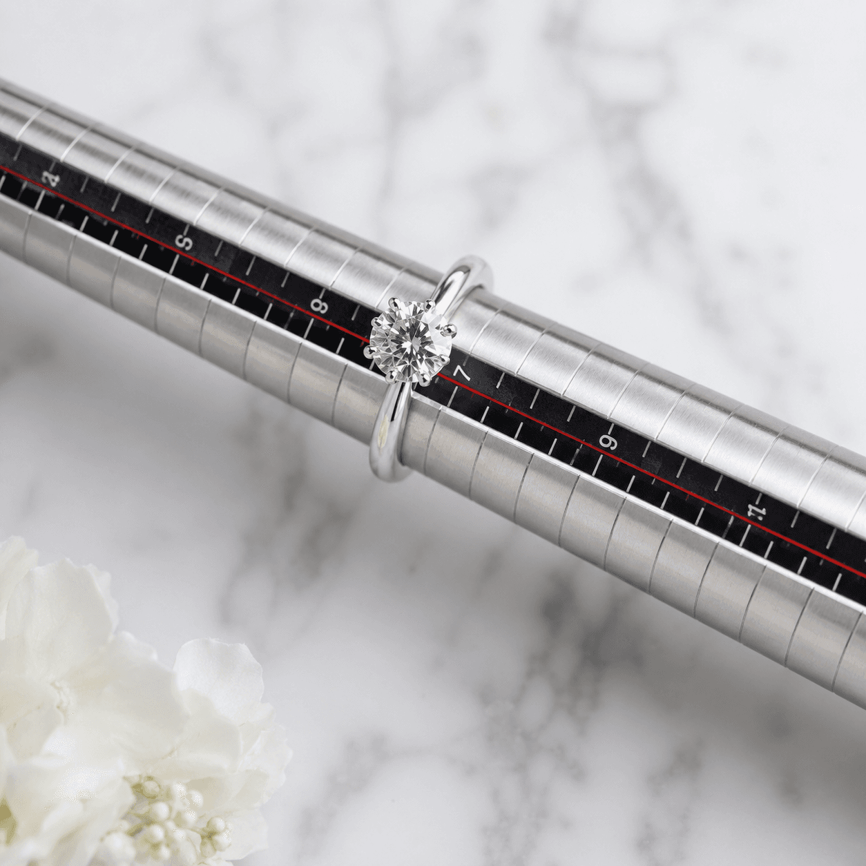 A polished diamond engagement ring resting on a ring sizing gauge tool, overhead flat-lay composition, soft natural light on
