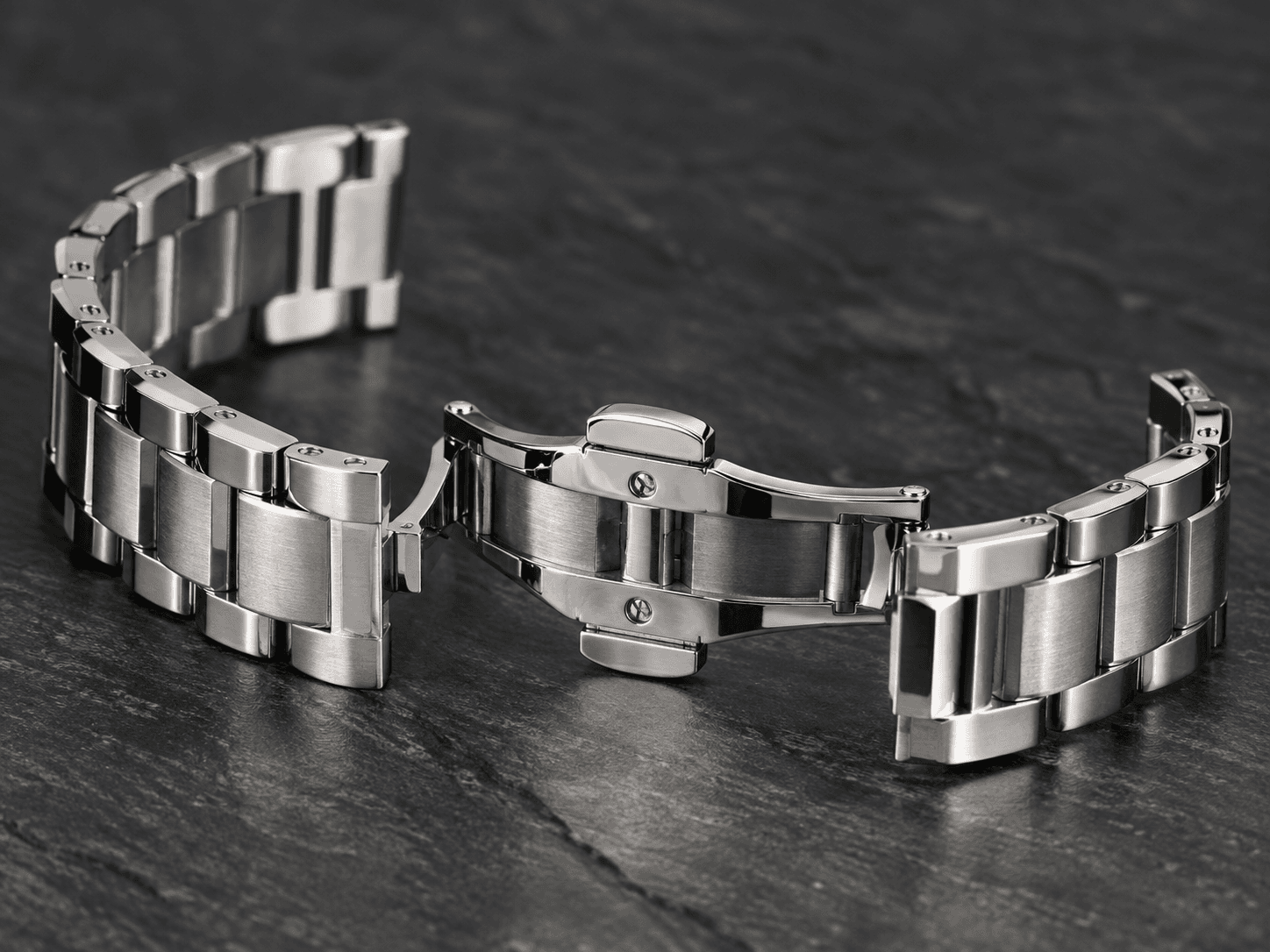 A macro shot of a stainless steel bracelet watch band with a butterfly clasp resting on a dark slate surface, studio