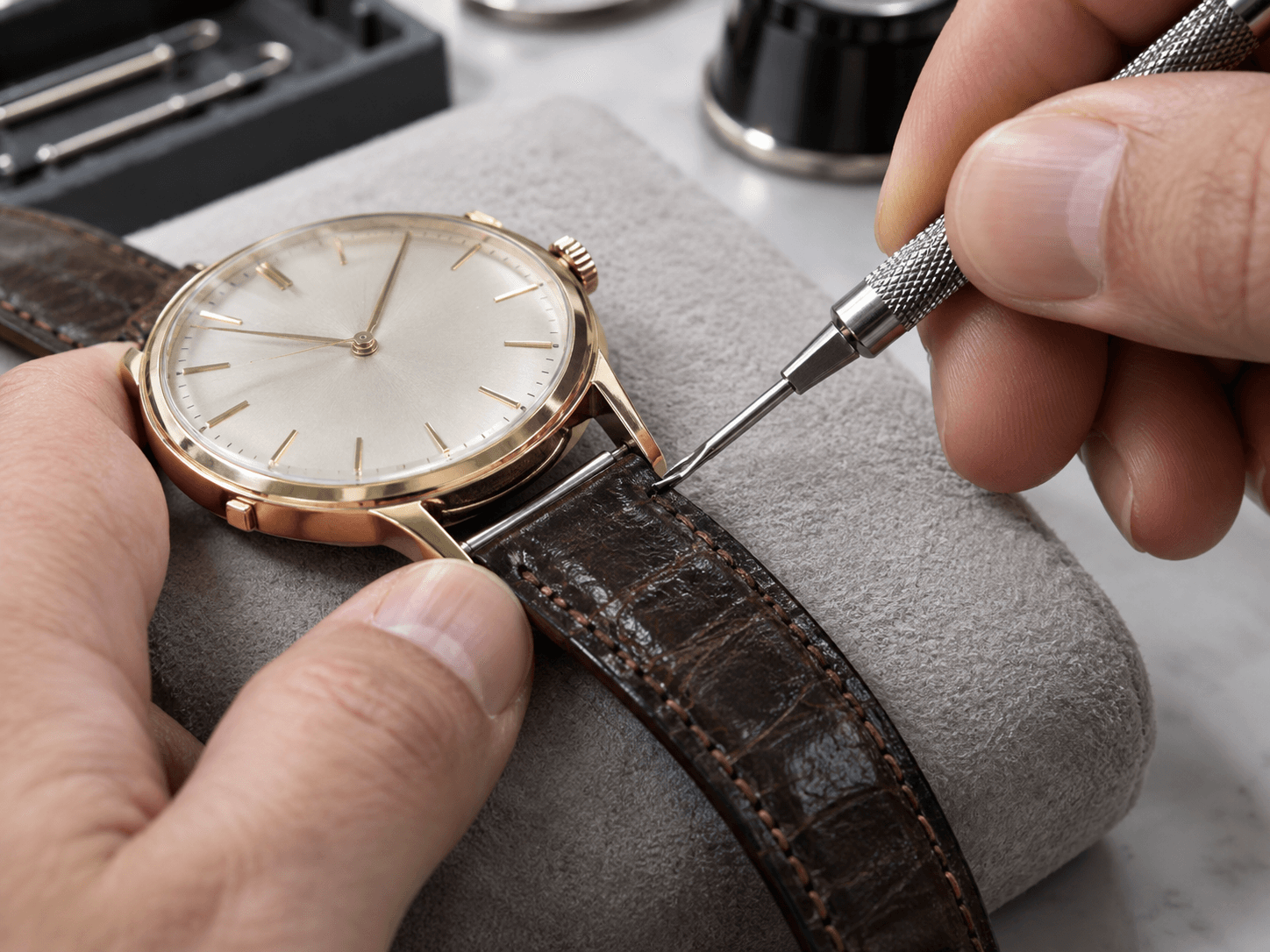 A jeweler using a spring bar tool to remove a worn watch band from a classic dress watch on a padded workbench, macro shot