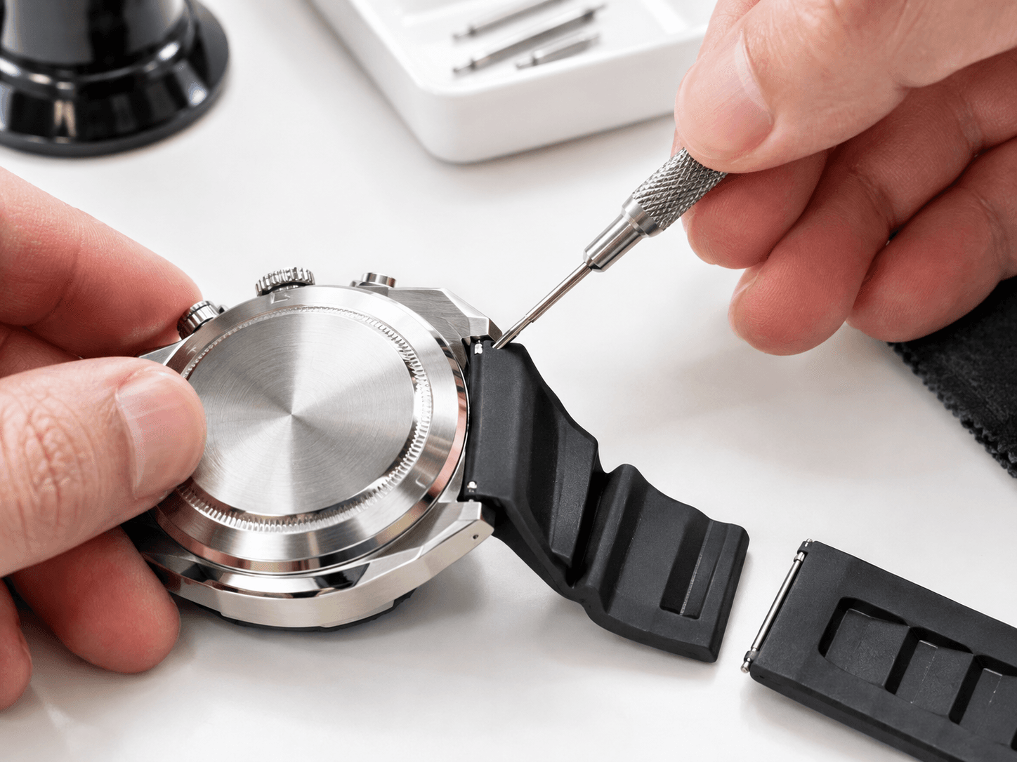 A jeweler using a spring bar tool to remove a rubber sport strap from a modern sports watch on a clean white workbench